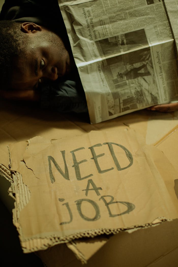 A man lies on cardboard indoors with a 'Need a Job' sign, highlighting social issues.