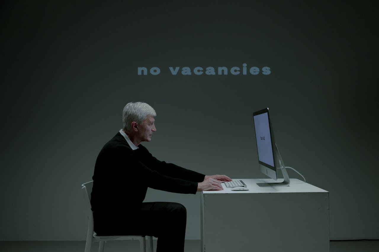 Elderly man using computer with 'no vacancies' displayed, highlighting job search struggles.